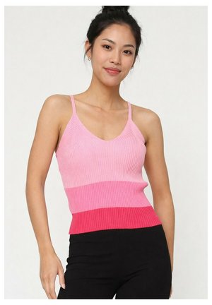 Femme aux cheveux foncés relevés en chignon, portant un débardeur côtelé dégradé rose et un pantalon noir, souriante sur un fond clair uni.