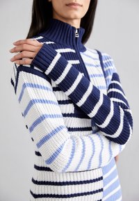 Pull en maille rayé bleu marine, bleu clair et blanc avec un col montant et une fermeture à moitié éclair. Caractéristiques des poignets côtelés contrastants et une texture travaillée.