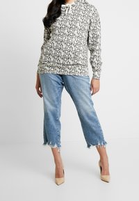 Šedá a černá vzorovaná mikina s kapucí a šňůrkovým límcem, doplněná o potrhané světle modré džíny a tělové vysoké podpatky.