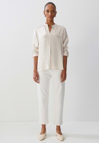 Blusa beige chiaro a maniche lunghe con scollatura aperta e polsini arrotolati, abbinata a pantaloni bianchi a gamba dritta e scarpe slip-on color crema.