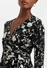 Czarna sukienka w kwiatowy wzór w białym kolorze, z długimi rękawami, dekoltem w serek i wiązaniem w talii. Gładki materiał o miękkiej teksturze.