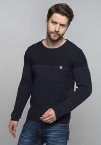 Pull en maille bleu marine avec un design texturé, présentant une section supérieure côtelée et un petit badge logo sur la poitrine. Associé avec un jean bleu.