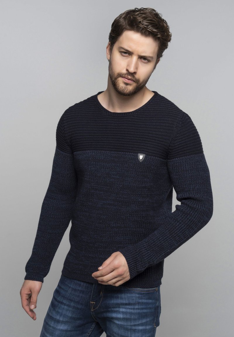 Pull en maille bleu marine avec un design texturé, présentant une section supérieure côtelée et un petit badge logo sur la poitrine. Associé avec un jean bleu.