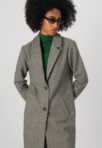 Pelēks un melns houndstooth raksta mētelis ar izgrieztu apkakli, divām priekšējām kabatām un brūniem pogām, virs zaļa turtleneck krekla.
