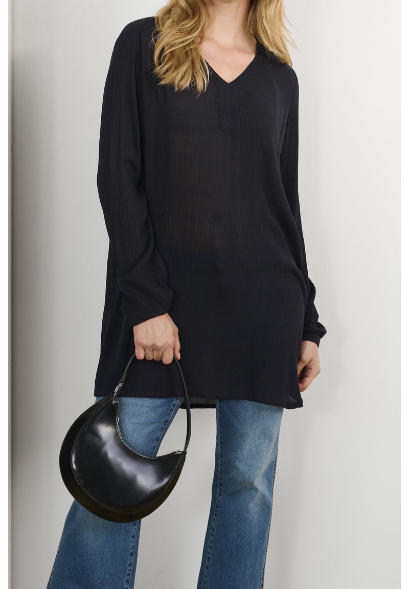 Femme portant un long chemisier noir et un jean bleu, tenant un sac à main noir en forme de croissant par sa poignée.