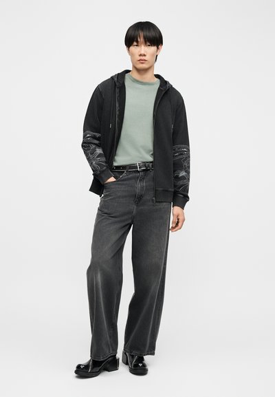 Νέος άνδρας που φοράει μαύρο φούτερ με σχέδια στα μανίκια, πράσινο μπλουζάκι, φαρδιά μαύρα τζιν και μαύρα παπούτσια με πλατφόρμα, στέκεται μπροστά σε ένα λευκό φόντο.