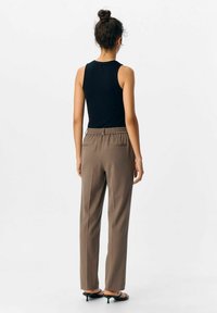 Femme de dos portant un haut sans manches noir, un pantalon taille haute marron et des sandales à talons noires sur un fond blanc.