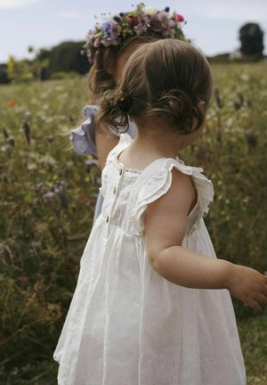 Robe blanche à volants avec des boutons et une broderie florale. Tissu doux et léger. Le bras d'un enfant est visible, jouant dans un champ.