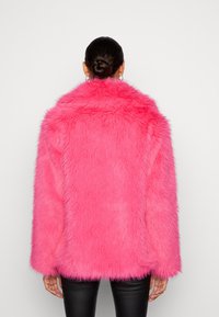 Różowa kurtka z sztucznego futra z szerokim kołnierzem, oversize'owy fason, długie rękawy i teksturowana powierzchnia. Nosi na sobie czarne dopasowane spodnie.