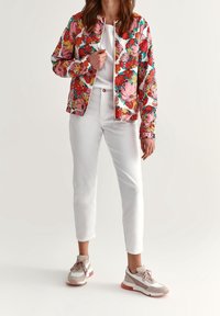 Blomstret bomberjakke i klare farver, hvid base med store pink og orange blomster, kombineret med hvide tætsiddende bukser og multicolor sneakers.