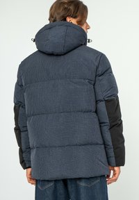 Námořnická puffer bunda s kostkovaným vzorem, prošívanou texturou a černými akcenty na rukávech. Obsahuje kapuci a má uvolněný střih.