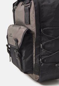 Sac à dos noir et gris avec une finition texturée, doté d'une poche frontale, de bretelles ajustables et de cordons décoratifs sur le côté.