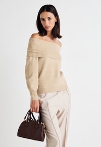 Beige off-the-shoulder sweater with ribbed neckline and long sleeves, paired with light trousers and a small brown handbag.