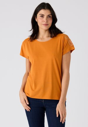 Frau mit dunklem Haar, die ein orangefarbenes kurzärmeliges Oberteil mit Spitzen-Details an den Schultern und dunkelblaue Jeans trägt, steht vor einem schlichten Hintergrund.