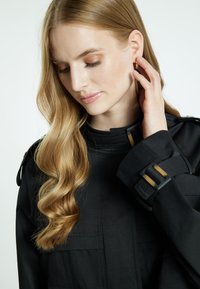 Zwarte, op maat gemaakte jas met een hoge kraag, voorzien van goudkleurige accenten en gestructureerde mouwen. Gladde textuur en moderne ontwerpelementen.