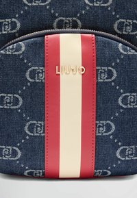 Zaino in denim con motivo a logo, striscia verticale rossa e bianca e un emblema dorato "LIUJO" sulla tasca frontale. Chiusura a zip.