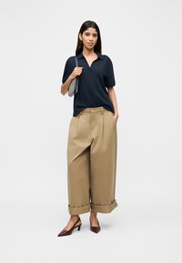 Μπλε μπλουζάκι πόλο με απαλό ύφασμα, άνετο μπεζ παντελόνι με φαρδιά μπατζάκια και μπορντό γόβες με μυτερή μύτη, κρατώντας ένα τσαντάκι με σχέδια.