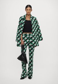 Cappotto e pantaloni con motivo floreale verde, abbinati a una maglietta nera. L'outfit ha una finitura lucida, accompagnato da una borsa nera.