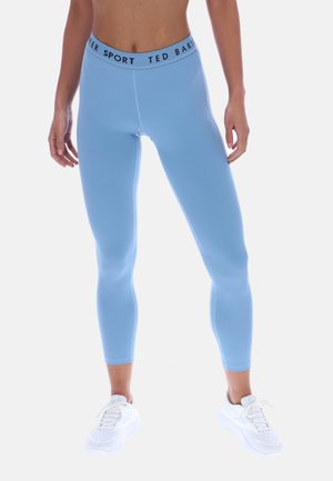 Kvinne iført lyseblå cropped leggings med "Ted Baker Sport" midjebelte og hvite treningssko, stående mot en ensfarget bakgrunn.