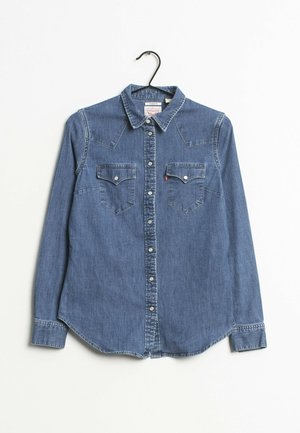 Chemise en denim bleu à manches longues avec boutons, deux poches poitrine, col plié et boutons-pression, suspendue sur un cintre noir sur fond blanc.