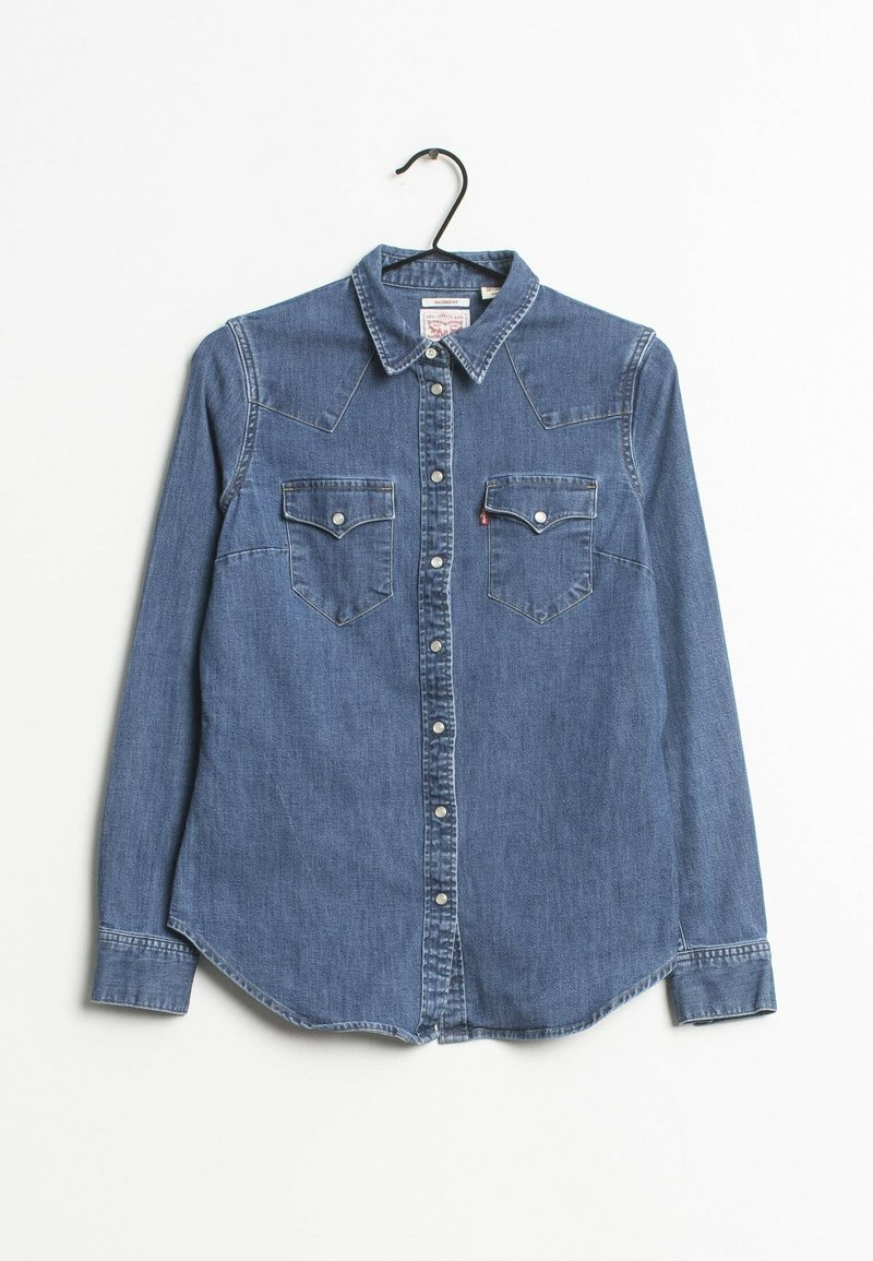 Chemise en denim bleu à manches longues avec boutons, deux poches poitrine, col plié et boutons-pression, suspendue sur un cintre noir sur fond blanc.