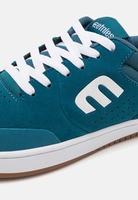 Teal suede sneaker features white laces and a white logo. The design includes perforations for ventilation and a gum rubber sole.