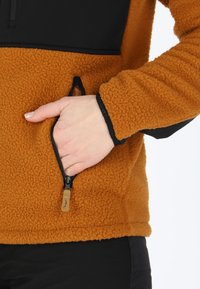 Giacca in pile di colore arancione bruciato con un pannello superiore nero. Presenta una tasca con zip e polsini a costine per una vestibilità aderente. Texture morbida.