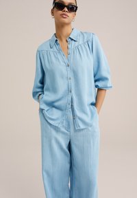 Helles blaues Denimhemd mit durchgeknöpftem Vorderteil, kurzen Ärmeln und abgerundetem Saum. Kombiniert mit passenden weiten Hose. Glatte Textur.