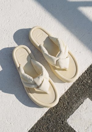 Par cremefarvede polstrede flip-flop sandaler, der hviler på tekstureret hvidt og sort fortov i sollys.