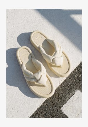 Egy pár krémszínű párnázott flip-flop szandál pihen texturált fehér és fekete járdán napsütésben.