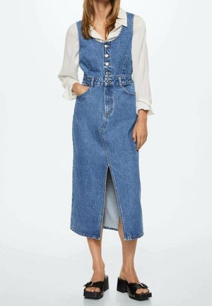 Kvinde iført en knappet blå denim midi-kjole over en hvid bluse med lange ærmer og sorte slip-on platform-sandaler.