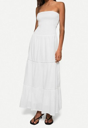 Kobieta w białej, warstwowej sukience maxi bez ramiączek z przylegającym karczkiem i czarnych sandałkach na obcasie, stojąca na jednolitym tle.