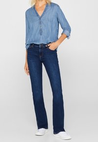 Jasnoniebieska koszula z chambray z dekoltem w serek, z detalami w postaci guzików i podwiniętymi rękawami, zestawiona z ciemnoniebieskimi dżinsami w stylu dzwonów oraz białymi sneakersami.