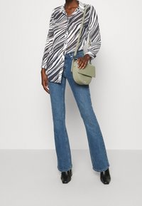 Donna che indossa una blusa a stampa zebrastripe bianco e nero, jeans blu, stivali neri con tacco e tiene in mano una borsa a tracolla verde chiaro.