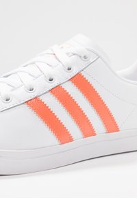 Vit lädersneaker med tre texturerade orange ränder på sidan och vita snören, visad i närbild.