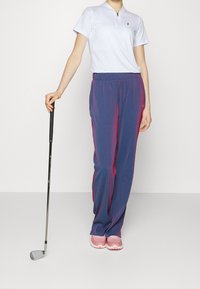 Tenue de golf avec un polo zippé blanc et un pantalon rayé bleu accentué de rose. Chaussures roses, tenant un club de golf. Design épuré, coupe sportive.