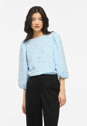 Nainen, jolla on olkapäähän ulottuvat tummat hiukset, yllään vaaleansininen, kuvioitu paita, jossa on pöyheät hihat, sekä mustat räätälöidyt housut.