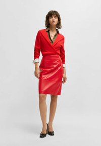 Gonna rossa in ecopelle con un design aderente e dettaglio drappeggiato davanti, abbinata a un top avvolgente rosso e una blusa verde oliva sotto.