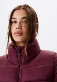 Bordeaux farvet puffjakke med høj krave og lynlås lukning. Har vandrette syninger og en rund trækknap. Glat, blank overflade.