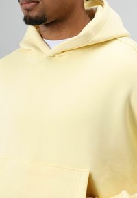 Lichtgele hoodie van zachte stof, voorzien van een kangoeroezak en een capuchon met trekkoord. Soepele textuur met geribbelde details.