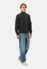 Giacca nera con zip e colletto alto, due tasche frontali e polsini a coste, abbinata a jeans larghi blu chiaro e sneakers crema.