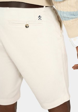 Primer plano de una persona que lleva pantalones cortos beige con un bolsillo trasero abotonado y un pequeño bordado de palos de golf cruzados cerca del pasador del cinturón.