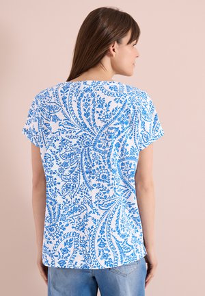 Femme portant un haut blanc à manches courtes avec un motif cachemire bleu complexe, dos tourné sur un fond beige uni.