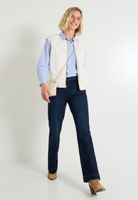 Gilet sans manches blanc et texturé par-dessus une chemise à boutons bleu rayée ; jeans bleu foncé ; bottes à talons beige ; tenant un petit sac tissé.