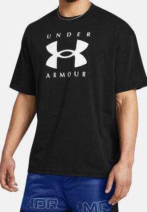 Mężczyzna noszący czarną koszulkę Under Armour z dużym białym logo i napisem oraz niebieskie sportowe szorty z siateczki.