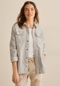 Giacca camicia in corduroy a coste di un grigio chiaro, con bottoni davanti, due tasche sul petto, maniche arrotolate e una consistenza morbida. Abbinata a pantaloni beige.
