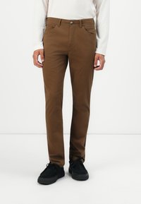 Man in een bruine slim-fit broek, witte lange mouw shirt en zwarte veterschoenen, staande tegen een witte achtergrond.