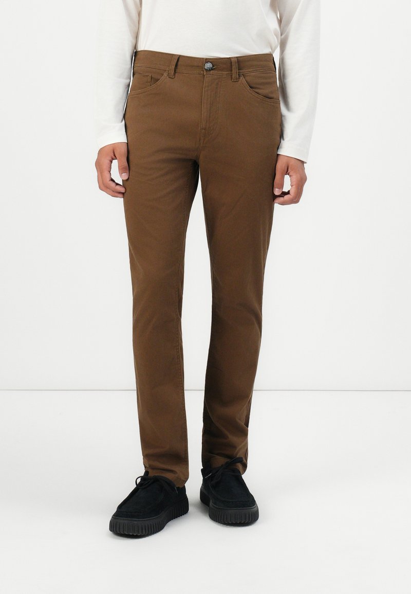 Man in een bruine slim-fit broek, witte lange mouw shirt en zwarte veterschoenen, staande tegen een witte achtergrond.