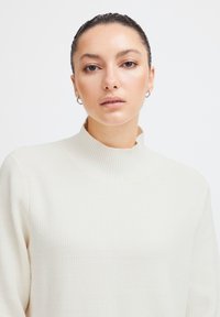 Pulover crem, cu guler înalt, din tricot cu model texturat și cu un croi lejer, așezat pe un fundal simplu.