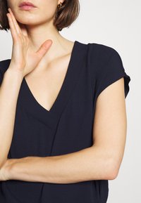 Top blu navy con scollo a V e maniche corte. Tessuto morbido e liscio; le cuciture si curvano attraverso il petto. Design semplice senza motivi o elementi hardware visibili.